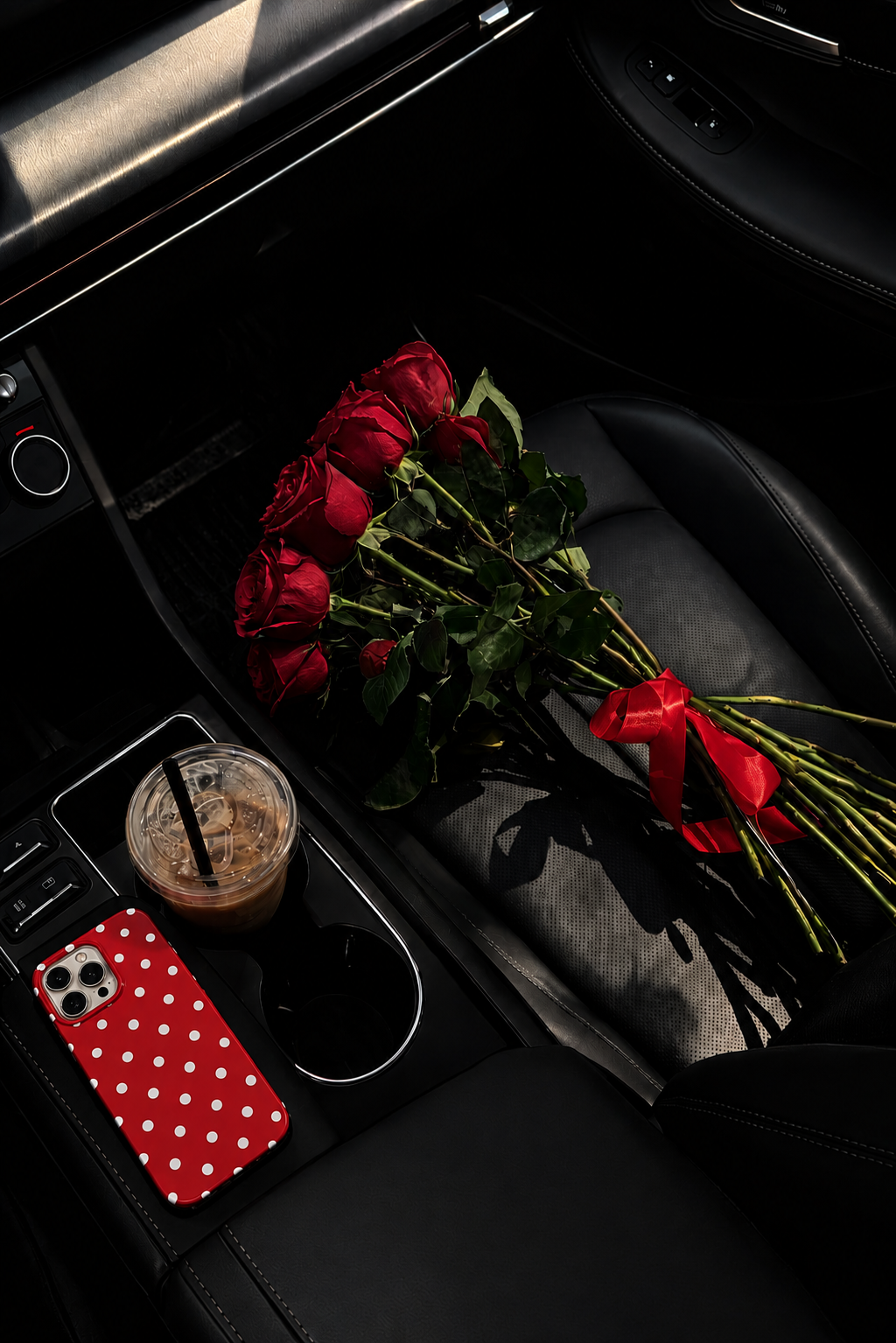 Bouquet of red roses on a passenger seat and a coffee cup with a straw plus a phone with a red and white polka dot on a car's center console.